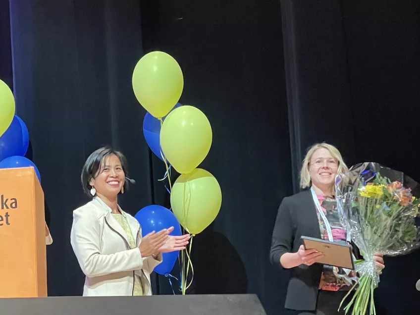 A woman receives a diploma and flowers, balloons in the background